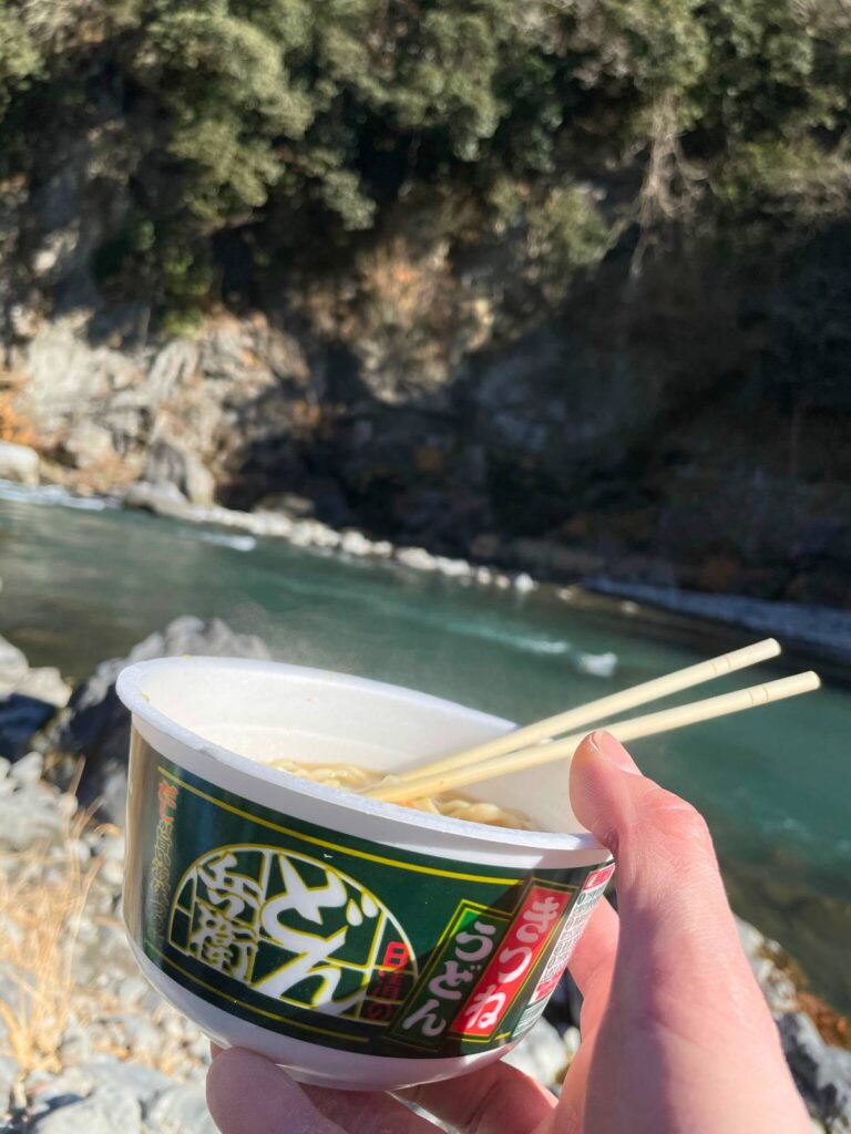 自然の景色を背景に、手に持ったカップ麺を外で楽しむ様子
