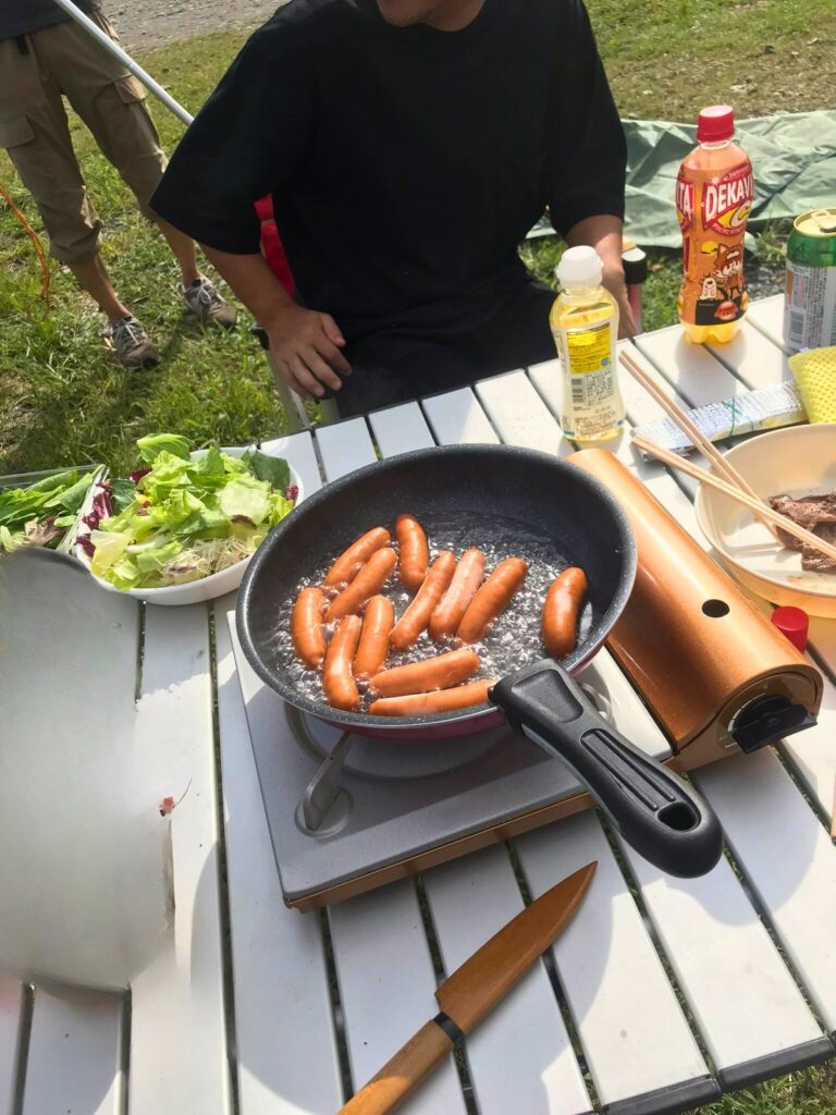 屋外でカセットコンロとフライパンを使ってソーセージを調理している様子